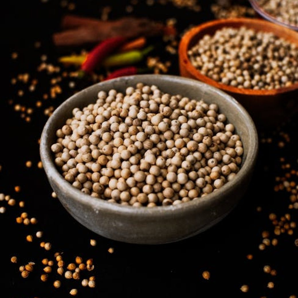 Close-up shot of white peppercorns in a ceramic bowl with scattered spices on dark background.